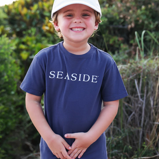 A smiling young boy wearing a navy blue t-shirt and a white baseball cap, both featuring the word 'SEASIDE' in white, standing outdoors in front of a blurred green background.