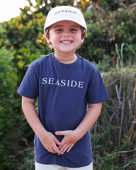 A smiling young boy wearing a navy blue t-shirt and a white baseball cap, both featuring the word 'SEASIDE' in white, standing outdoors in front of a blurred green background.