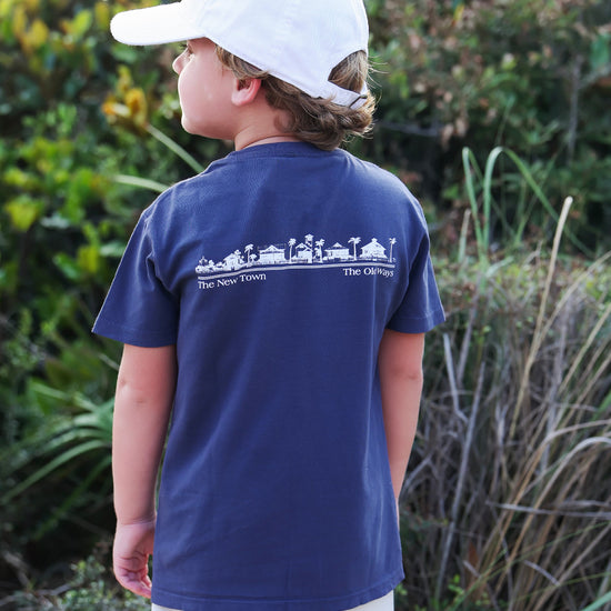 A child wearing a navy blue t-shirt with a white graphic print on the back, a white baseball cap, and light beige shorts, standing outdoors in front of green foliage.