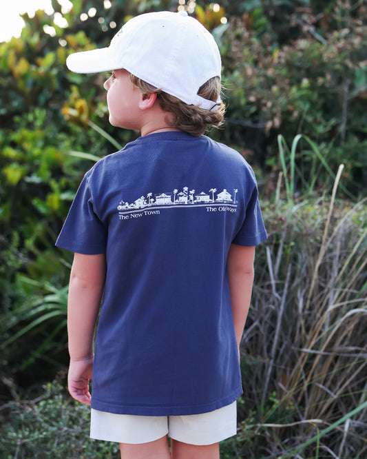 A child wearing a navy blue t-shirt with a white graphic print on the back, a white baseball cap, and light beige shorts, standing outdoors in front of green foliage.