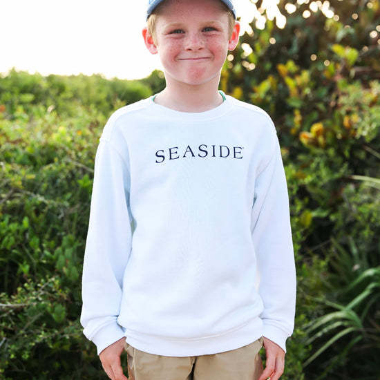A young boy wearing a white long-sleeve crewneck sweatshirt with 'SEASIDE' printed in navy blue across the chest, khaki shorts, and a light blue baseball cap, standing outdoors in front of a green bush.