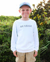 A young boy wearing a white long-sleeve crewneck sweatshirt with 'SEASIDE' printed in navy blue across the chest, khaki shorts, and a light blue baseball cap, standing outdoors in front of a green bush.