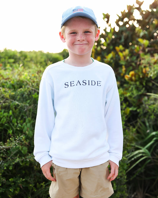 A young boy wearing a white long-sleeve crewneck sweatshirt with 'SEASIDE' printed in navy blue across the chest, khaki shorts, and a light blue baseball cap, standing outdoors in front of a green bush.