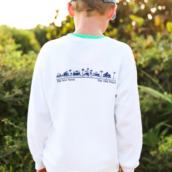 A child viewed from behind wearing a white crewneck sweatshirt with a blue graphic print on the back, khaki pants, and a light blue baseball cap, set against a blurred outdoor background.