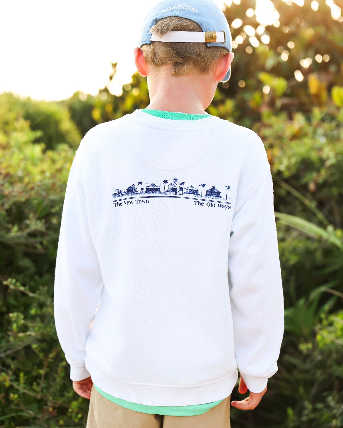 A child viewed from behind wearing a white crewneck sweatshirt with a blue graphic print on the back, khaki pants, and a light blue baseball cap, set against a blurred outdoor background.