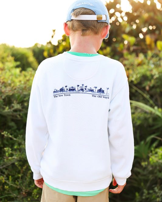 A child viewed from behind wearing a white crewneck sweatshirt with a blue graphic print on the back, khaki pants, and a light blue baseball cap, set against a blurred outdoor background.