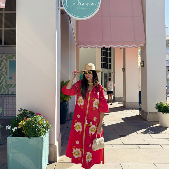 A woman stands on a sunny outdoor shopping walkway wearing a bright red midi dress with a white and yellow floral print, a beige straw hat, and carrying a small white handbag. A pink awning and a round teal sign reading 'Cabana' are visible in the background.