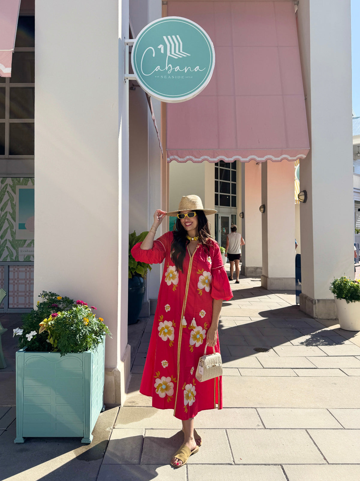 A woman stands on a sunny outdoor shopping walkway wearing a bright red midi dress with a white and yellow floral print, a beige straw hat, and carrying a small white handbag. A pink awning and a round teal sign reading 'Cabana' are visible in the background.