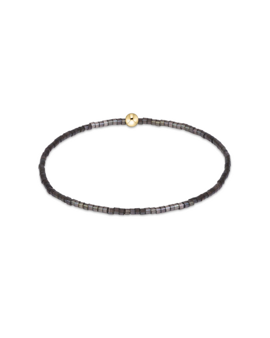 A delicate beaded stretch bracelet with small dark gray and black beads, featuring a single gold-toned round bead at the center, displayed on a clean white background.