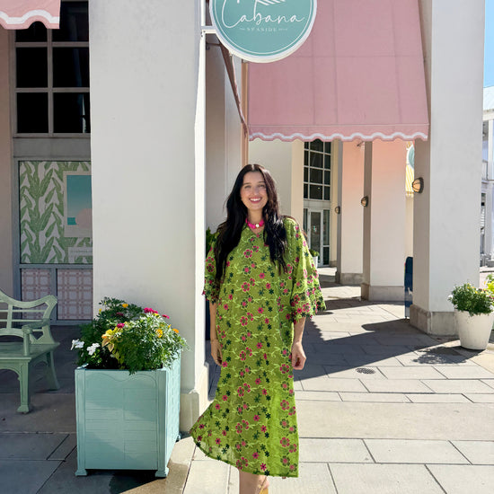 A woman stands on a sidewalk in front of a building with pink awnings and a round sign reading Cabana, wearing a bright green midi dress with a pink and red floral print and tan heeled sandals.