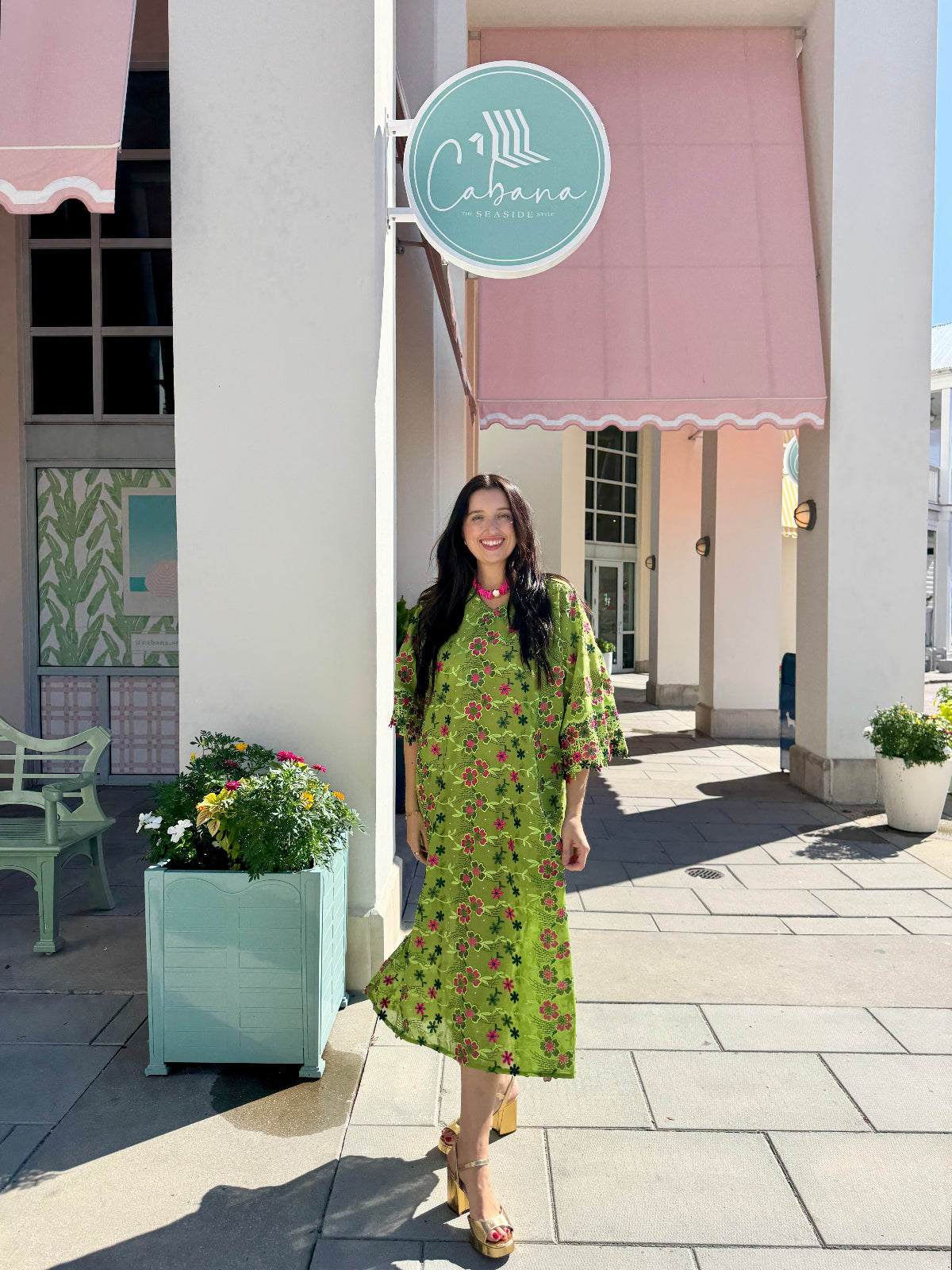 A woman stands on a sidewalk in front of a building with pink awnings and a round sign reading Cabana, wearing a bright green midi dress with a pink and red floral print and tan heeled sandals.