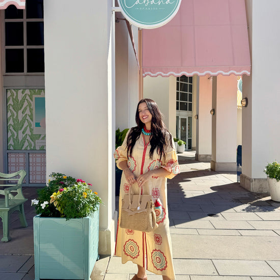 A woman stands on a sidewalk in front of a building with a pink awning and a round turquoise sign reading Cabana. She is wearing a long cream dress with colorful circular patterns, carrying a tan handbag and wearing brown sandals.