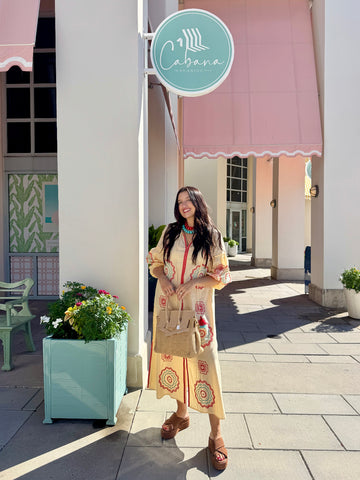 A woman stands on a sidewalk in front of a building with a pink awning and a round turquoise sign reading Cabana. She is wearing a long cream dress with colorful circular patterns, carrying a tan handbag and wearing brown sandals.