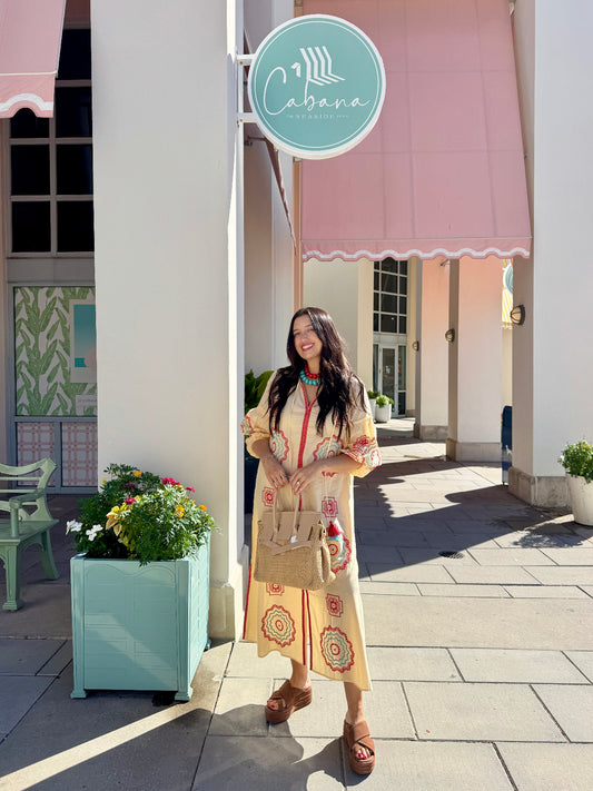 A woman stands on a sidewalk in front of a building with a pink awning and a round turquoise sign reading Cabana. She is wearing a long cream dress with colorful circular patterns, carrying a tan handbag and wearing brown sandals.