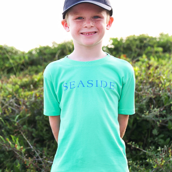 A smiling young boy wearing a bright mint green short-sleeve t-shirt with 'SEASIDE' printed in blue across the chest, paired with a navy blue baseball cap and khaki pants, standing outdoors in front of green bushes.
