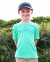 A smiling young boy wearing a bright mint green short-sleeve t-shirt with 'SEASIDE' printed in blue across the chest, paired with a navy blue baseball cap and khaki pants, standing outdoors in front of green bushes.