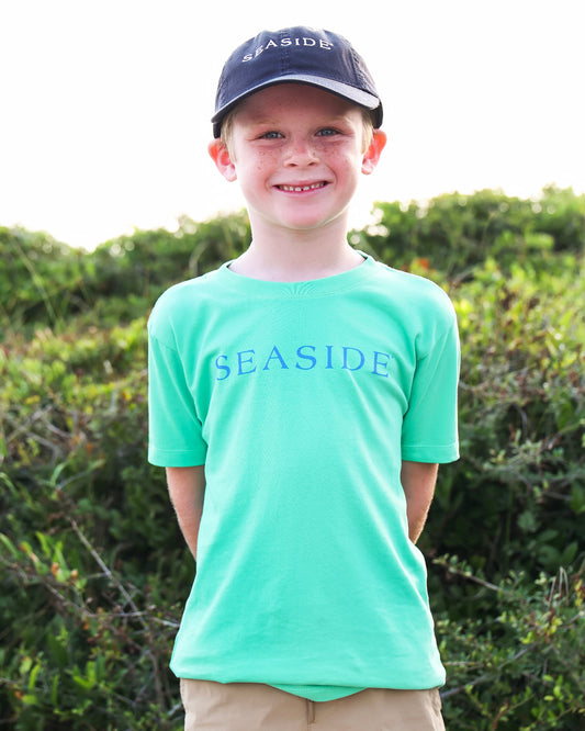 A smiling young boy wearing a bright mint green short-sleeve t-shirt with 'SEASIDE' printed in blue across the chest, paired with a navy blue baseball cap and khaki pants, standing outdoors in front of green bushes.
